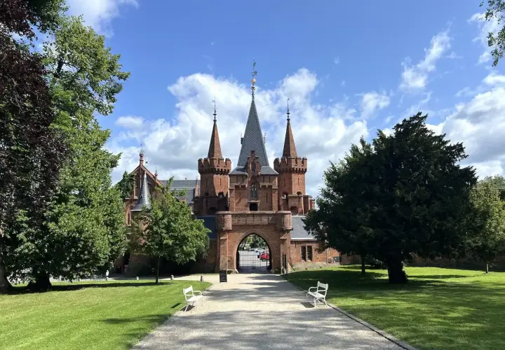 Imponujący, czerwony zamek z wieżyczkami i bramą wjazdową, otoczony zielonym parkiem i drzewami.