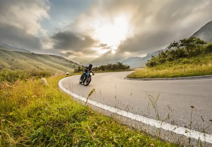 Un motociclista affronta una curva su una strada di montagna, godendosi un perfetto weekend in moto tra paesaggi mozzafiato e cielo nuvoloso.