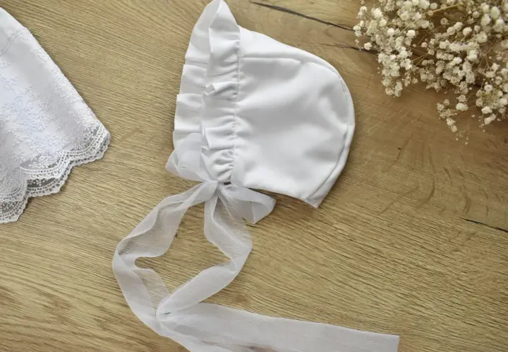 Elegancka, biała bonetka do chrztu z tiulową kokardką i koronkowym wykończeniem.