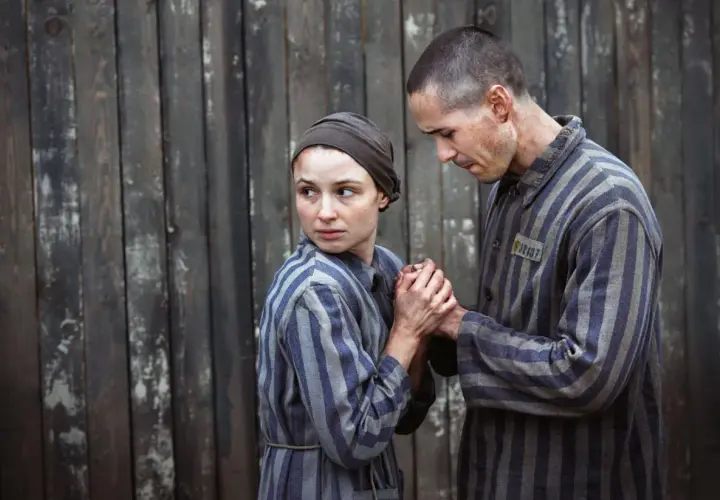 Para więźniów w pasiakach, scena z filmu o Auschwitz. Trzymają się za ręce, patrząc na siebie z nadzieją i strachem.