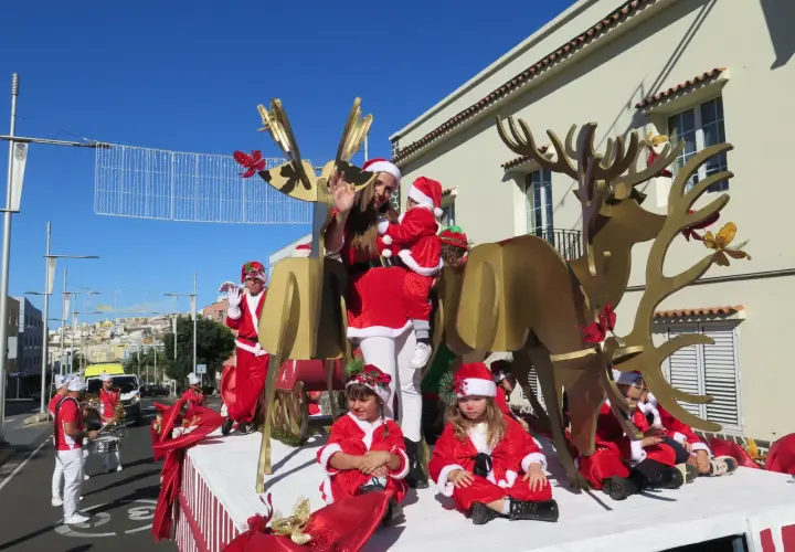 Niños y adultos disfrazados de Papá Noel y elfos participan en una cabalgata de Papá Noel, con un gran reno dorado y decoraciones navideñas.