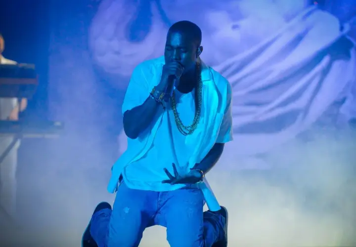 Kanye West na koncercie, klęczy z mikrofonem, otoczony niebieskim światłem i mgłą.