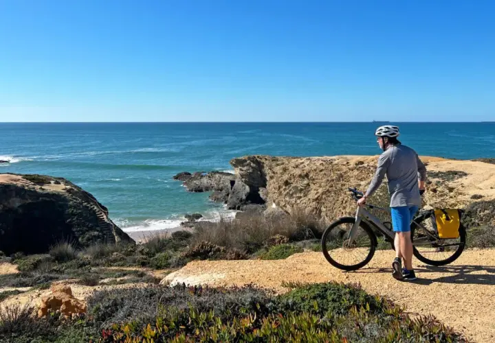 A cyclist pauses on a cliffside path during a Porto to Lisbon bike tour, admiring the vast blue ocean.