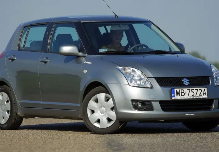 Szary Suzuki Swift 2007 z kierowcą za kierownicą, zaparkowany na drodze.
