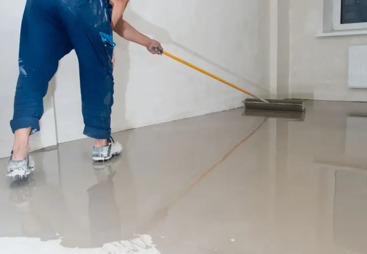Pracownik w niebieskich spodniach i butach z kolcami rozprowadza wałkiem świeżą wylewkę betonową. Sprawdź wylewka betonowa cena za m2 robocizny bez materiału.