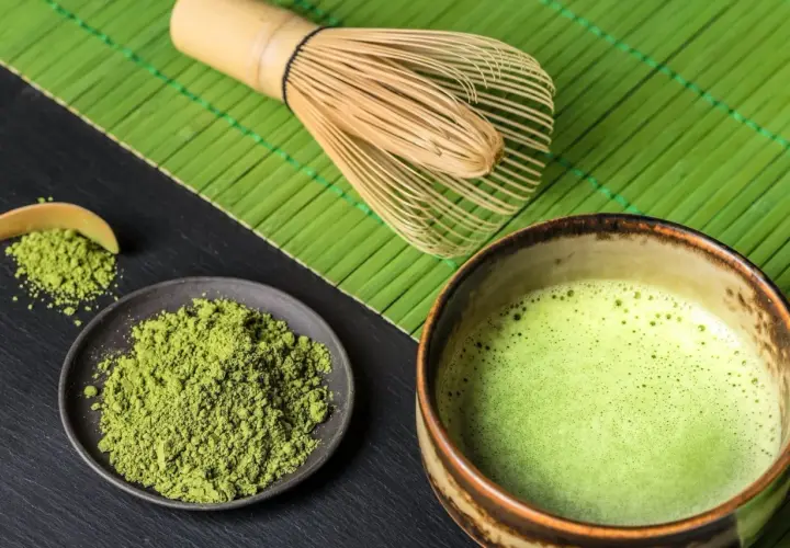 Matcha-Pulver auf einem Teller, daneben eine Schale mit schaumiger Matcha-Creme und ein Bambusbesen.