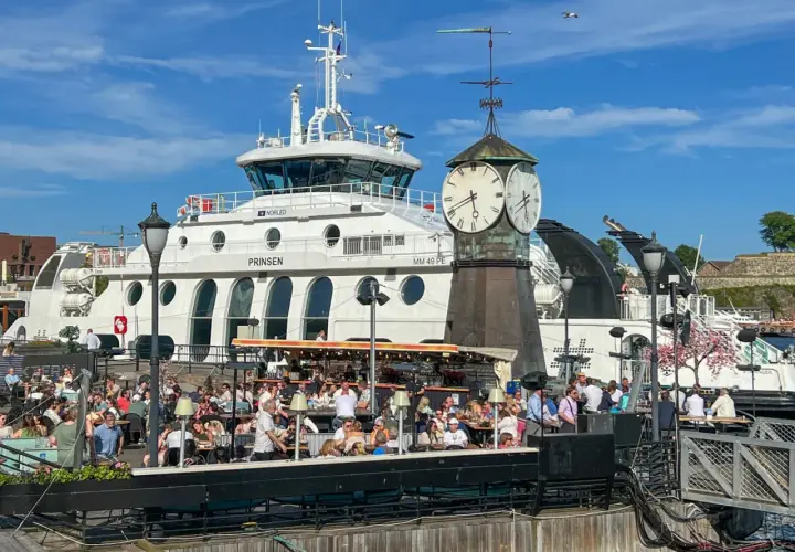 Tłum ludzi w kawiarni nad wodą w Oslo, z promem "Prinsen" i zegarem w tle. Ceny w Oslo mogą być wysokie, ale widok jest wart swojej ceny.