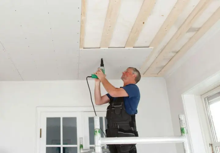 Man på byggställning skruvar gipsskivor i taket. Taket är under renovering med synliga träreglar.
