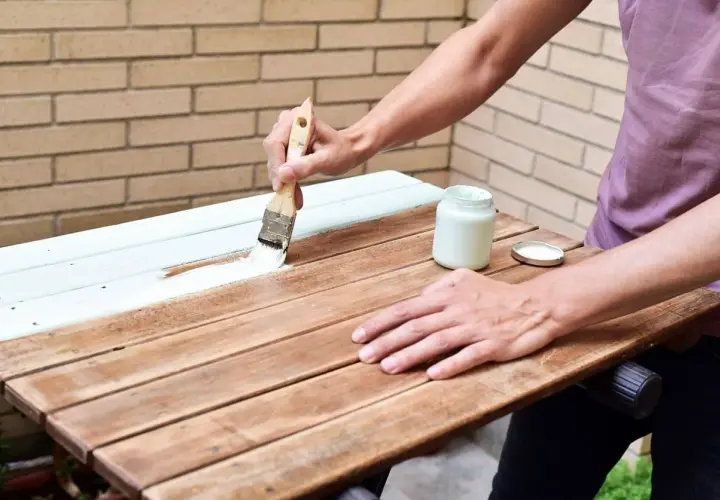 Persona pintando una mesa de madera exterior con brocha y pintura blanca.