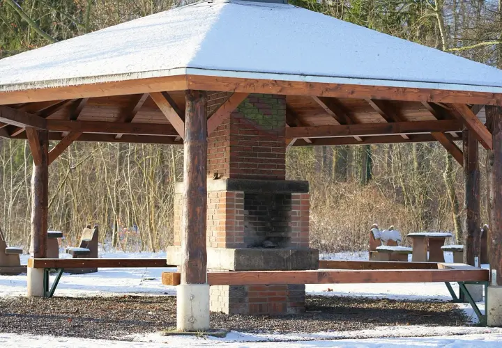 En träpaviljong med snö på taket och en murad grill i mitten. Bänkar och bord står redo för en mysig fika utomhus.