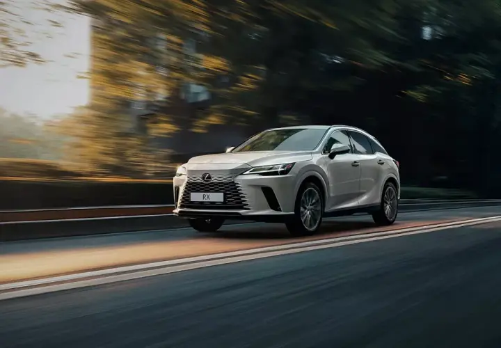Biały Lexus RX 350h pędzi po drodze, otoczony rozmazanymi drzewami i światłem zachodzącego słońca.