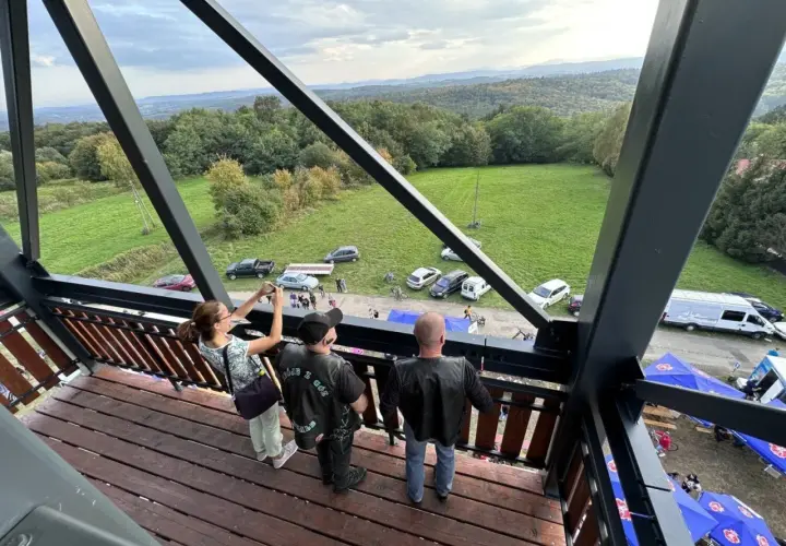 Widok z wieży widokowej na malowniczy krajobraz z lasami i polami. Na dole widać zaparkowane samochody, namioty i ludzi, jakby szykowali się na festyn lub zlot. Może to być nawet zlot motocyklowy, bo jeden z panów ma na sobie kamizelkę z napisem "GÓRY ...