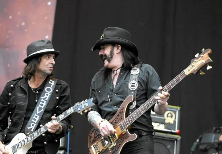 Lemmy Kilmister z zespołem na scenie. Dwóch muzyków gra na gitarach, jeden w kapeluszu, drugi w kowbojskim.
