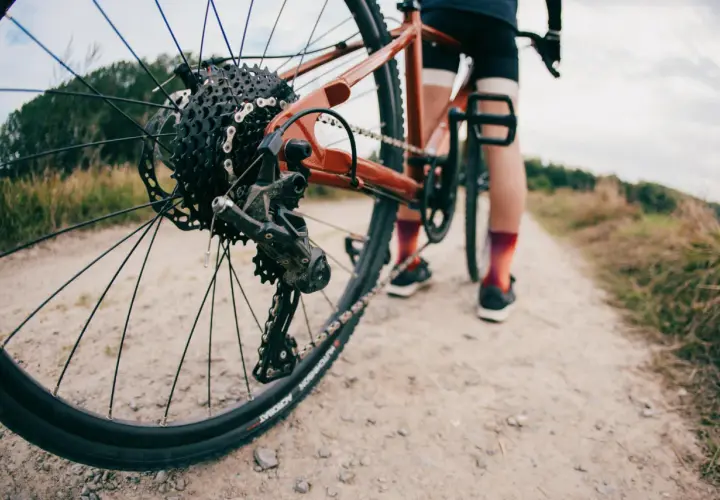 Widok z bliska tylnej przerzutki rowerowej, która wygląda na Shimano Acera lub Alivio, na tle szutrowej drogi i rowerzysty.