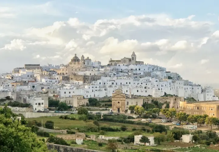 A white city perched on a hill, reminiscent of a scene from Puglia, Italy, with a church and scattered buildings.