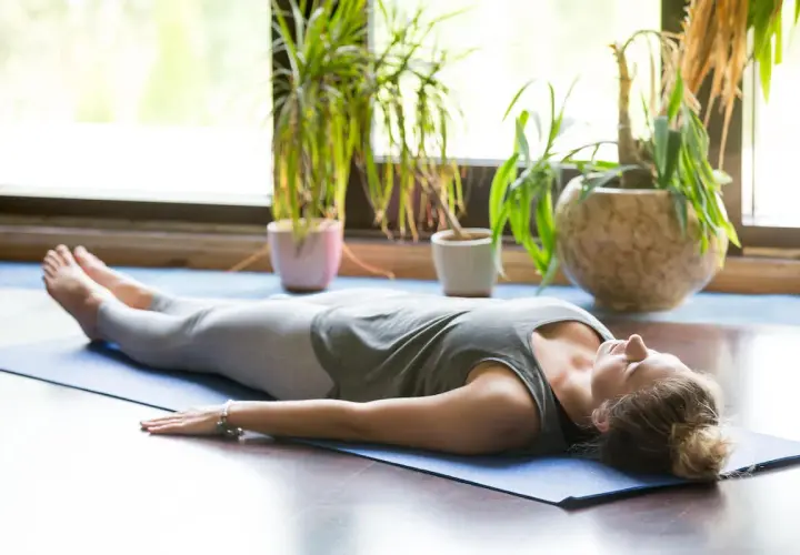 Kobieta w pozycji savasana, zrelaksowana na macie do jogi, otoczona zielenią roślin.