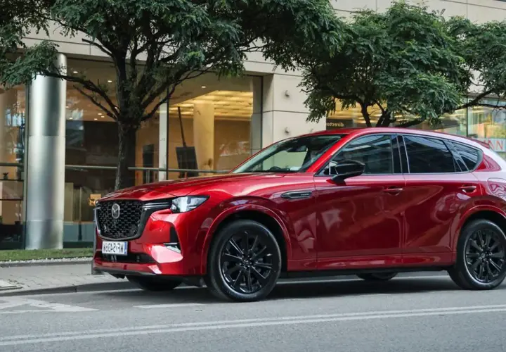 Lśniąca, czerwona Mazda CX-60 3.3 diesel na tle nowoczesnego budynku i drzew.