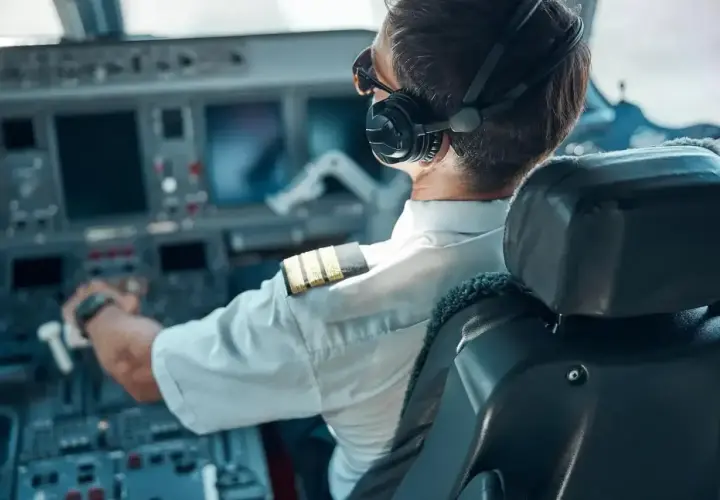 Pilot samolotu w słuchawkach i okularach przeciwsłonecznych, z ręką na sterach, w kabinie pełnej ekranów.