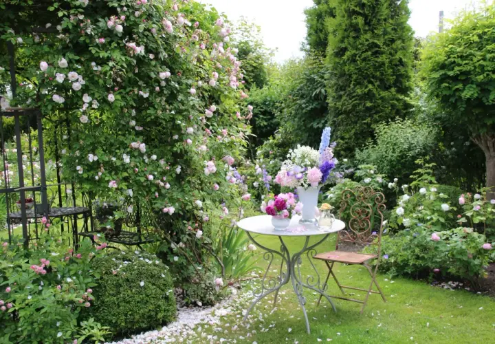 Ein romantischer Garten mit blühenden Rosen, ein Tisch mit Blumenarrangements und ein schmiedeeiserner Stuhl laden zum Verweilen ein. Ein idyllischer Ort, um einen **beet anlegen**-Traum zu verwirklichen.