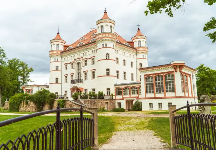 Zamek w Łomnicy, biała budowla z czerwonymi dachami i wieżyczkami, otoczona zielenią.