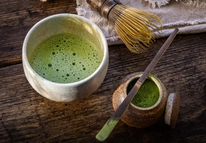 Un bol de matcha mousseux, une cuillère en bambou et de la poudre verte dans un pot sur une table en bois.