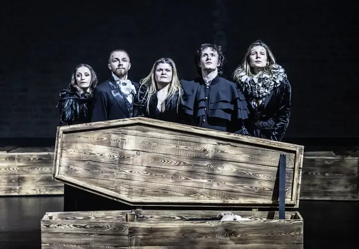 Aktorzy Teatru Pantomimy Wrocław w strojach z epoki stoją nad otwartą trumną, z której wystaje ręka.