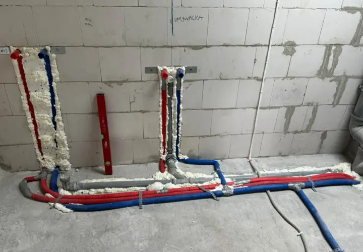 Postępująca budowa kanalizacji i instalacji wodnej w łazience. Rury wodne (czerwone i niebieskie) oraz odpływowe są częściowo obudowane pianką montażową.