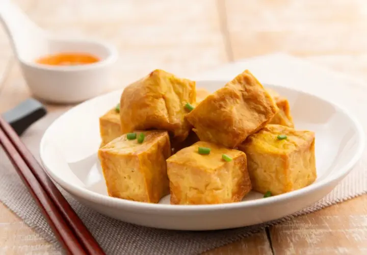 Tofu crujiente dorado, cortado en cubos y adornado con cebollín, servido en un plato blanco con salsa y palillos.