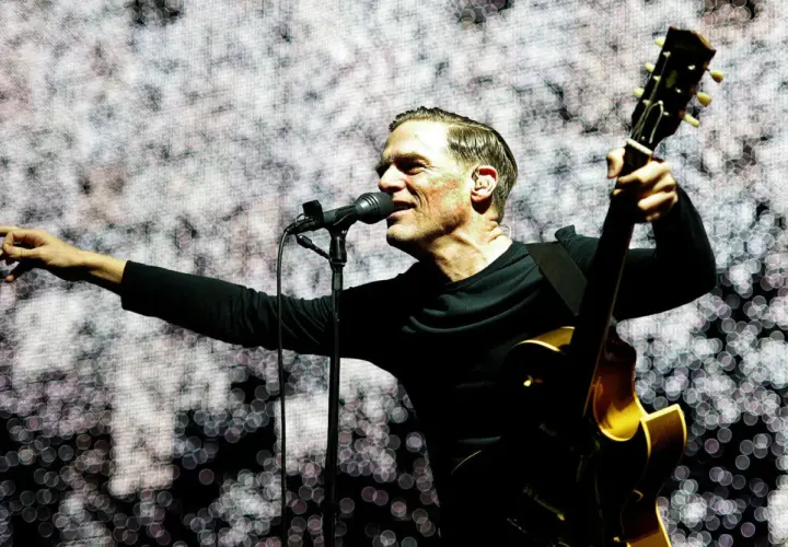 Bryan Adams na koncercie, śpiewa do mikrofonu i gra na gitarze.