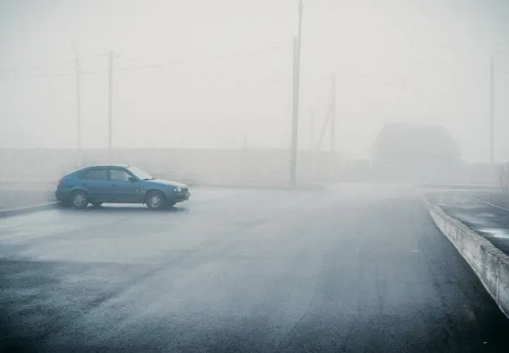 Un coche azul solitario se detiene en medio de una **niebla espesa** que oculta un camino y una casa.