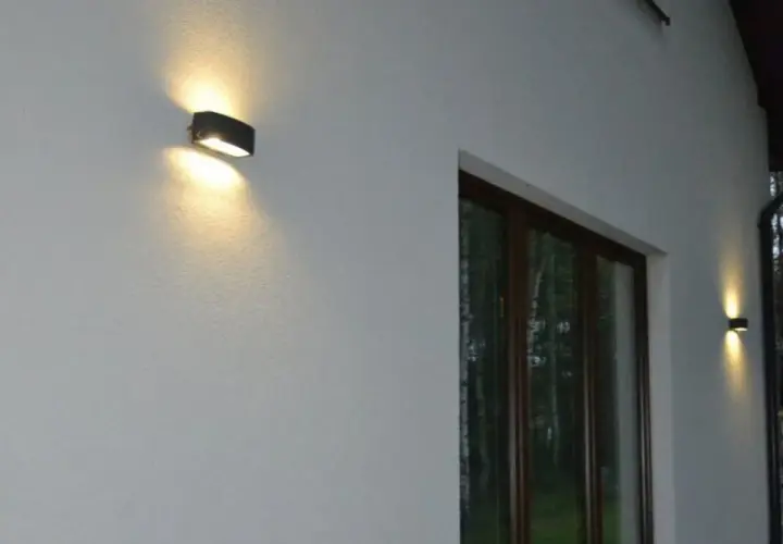 Nierówności na elewacji pod światło. Dwa kinkietowe punkty świetlne oświetlają białą, chropowatą ścianę domu, tworząc subtelne cienie.