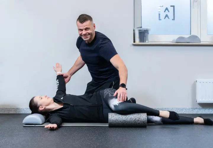 Fizjoterapeuta pomaga pacjentce w ćwiczeniach na kręgosłup. Kobieta leży na macie, z nogami ułożonymi na wałku, a mężczyzna koryguje jej pozycję ramienia.