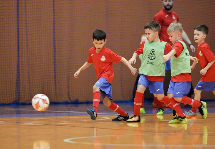Zacięte współzawodnictwo sportowe dzieci i młodzieży na hali. Mali piłkarze w czerwonych koszulkach i zielonych kamizelkach walczą o piłkę.
