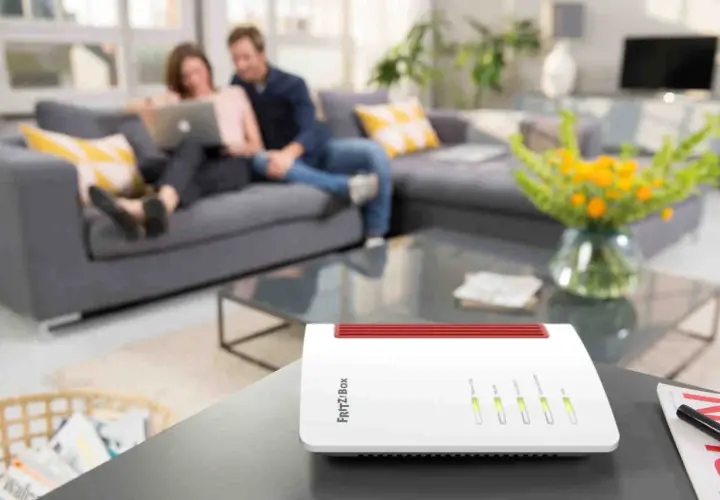 Biały router FRITZ!Box na stoliku, z parą relaksującą się na kanapie w tle. To urządzenie zapewnia domowe Wi-Fi.