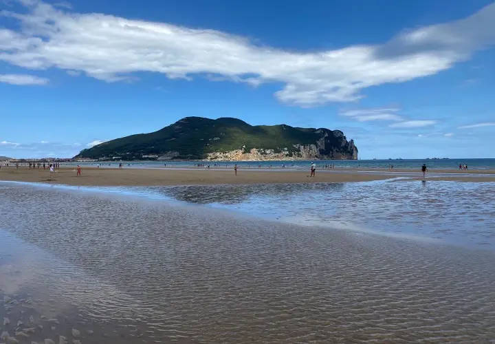 Amplia **playa Laredo** con gente paseando y jugando, bajo un cielo azul con nubes blancas y una gran montaña verde al fondo.