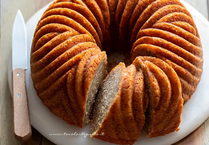 Una torta cachi e mele dorata, con un bordo scanalato, è affettata su un tagliere bianco. Un coltello con manico in legno è appoggiato accanto.