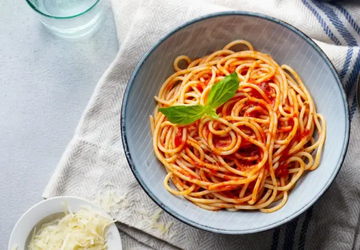 Spaghetti z sosem pomidorowym i bazylią w misce, obok starty ser i sztućce.