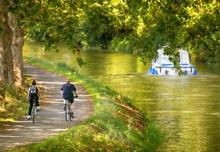 Pareja en bicicleta disfruta de un **canal du midi recorrido** bajo árboles frondosos, mientras una barcaza navega por el agua.