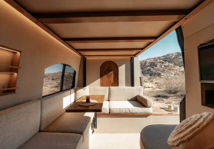 Intérieur de camping-car aménagé avec banquettes, table et porte en bois, vue sur un paysage désertique.