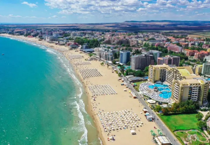 Słoneczna riwiera bułgarska z szeroką, piaszczystą plażą pełną leżaków i parasoli, turkusowym morzem i hotelami w tle.