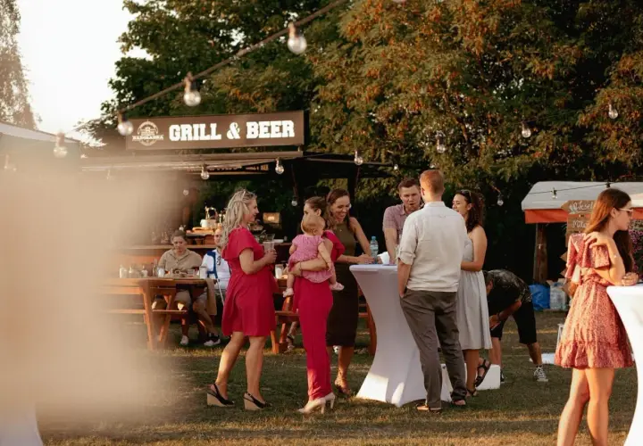 Ludzie rozmawiający przy stolikach i budce z napisem "Grill & Beer" na zewnątrz.