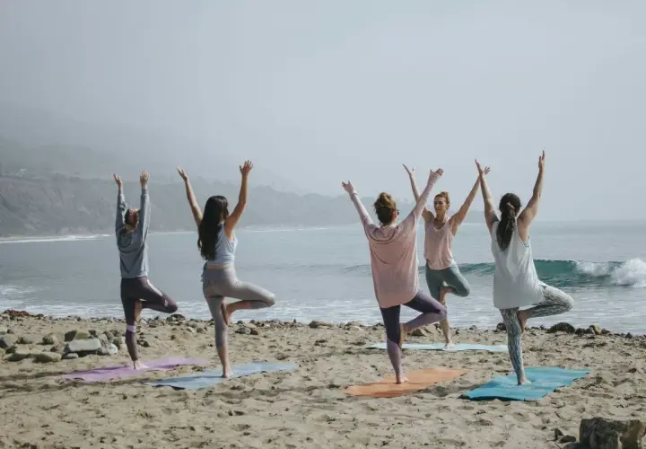 Fem kvinnor utför pilates yoga på stranden med havet i bakgrunden.