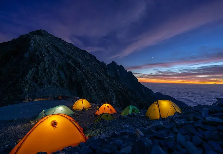 Acampada en la montaña al amanecer. Tiendas iluminadas sobre rocas, con un mar de nubes y cielo anaranjado. ¡Espero que no haya multa por acampar aquí!