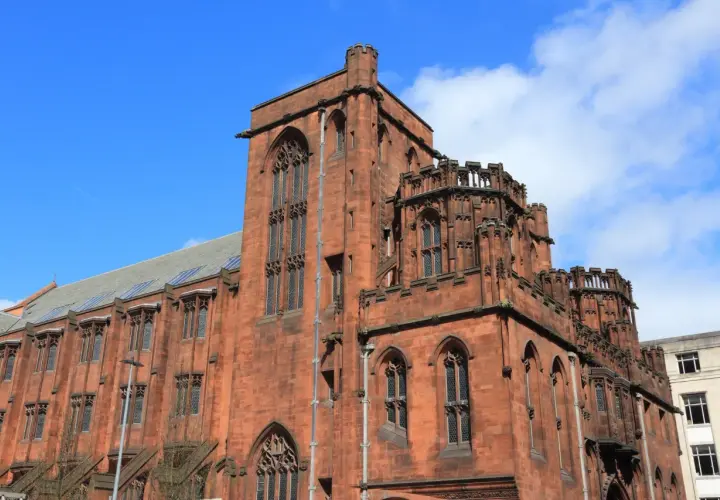 Imponująca, ceglana architektura w stylu gotyckim w Manchesterze, z charakterystycznymi wieżyczkami i łukowymi oknami.