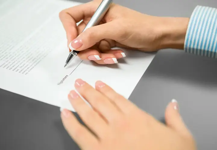 Mano femenina con uñas cuidadas escribe en un documento. El texto "Signature" es visible, pero el **contrato sin firmar** carece de **validez** hasta que se complete.