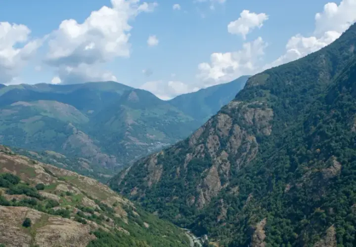 Paisaje montañoso con el bosque de Carlac cubriendo las laderas. Nubes blancas flotan en un cielo azul claro.