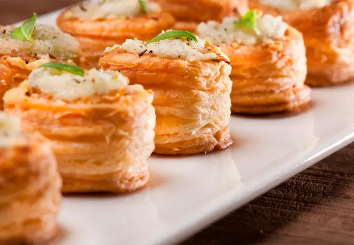 Petites bouchées à la reine dorées, garnies d'une préparation crémeuse et d'une feuille de basilic, présentées sur un plat blanc.