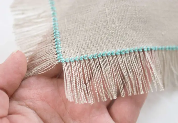Close-up of a hand holding linen fabric with a turquoise stitched edge, showing how to sew fringe.