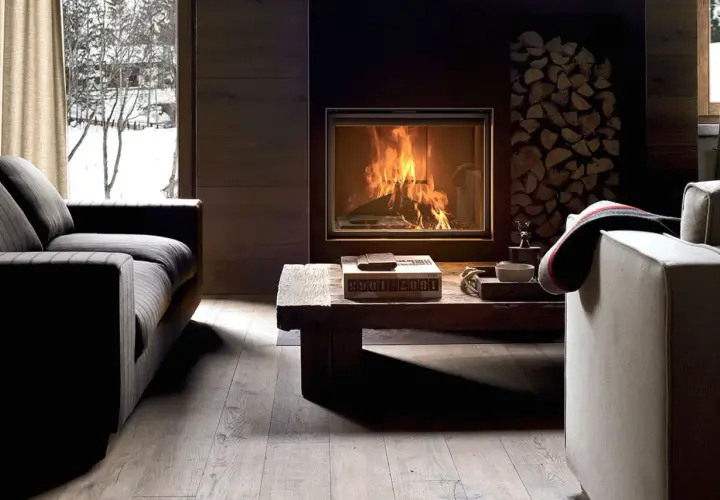 Gemütliches Wohnzimmer mit Kaminfeuer, Holzstapel und Blick auf verschneite Bäume. Tolle Kamin Ideen für Ihr Wohnzimmer.
