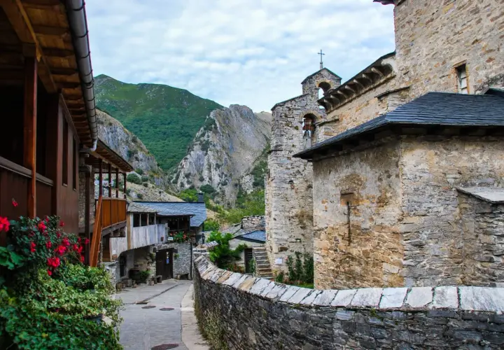 Un pueblo de piedra y madera se alza en el valle del silencio, rodeado de montañas verdes y un cielo nublado.